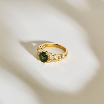 La Bague Saphir Teal • 2,07ct de Bonnot Paris présente une pierre précieuse verte en forme de cœur bordée de petites pierres claires, magnifiquement sertie sur un fond neutre doucement éclairé par des ombres douces.