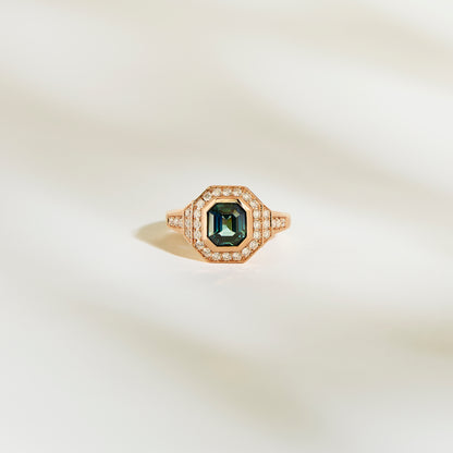 La Bague Saphir Teal de Bonnot Paris, une bague en or ornée d'une pierre précieuse vert foncé taille émeraude de 1,56 ct entourée d'un halo de petits diamants blancs, est élégamment mise en valeur sur un fond doux et clair.