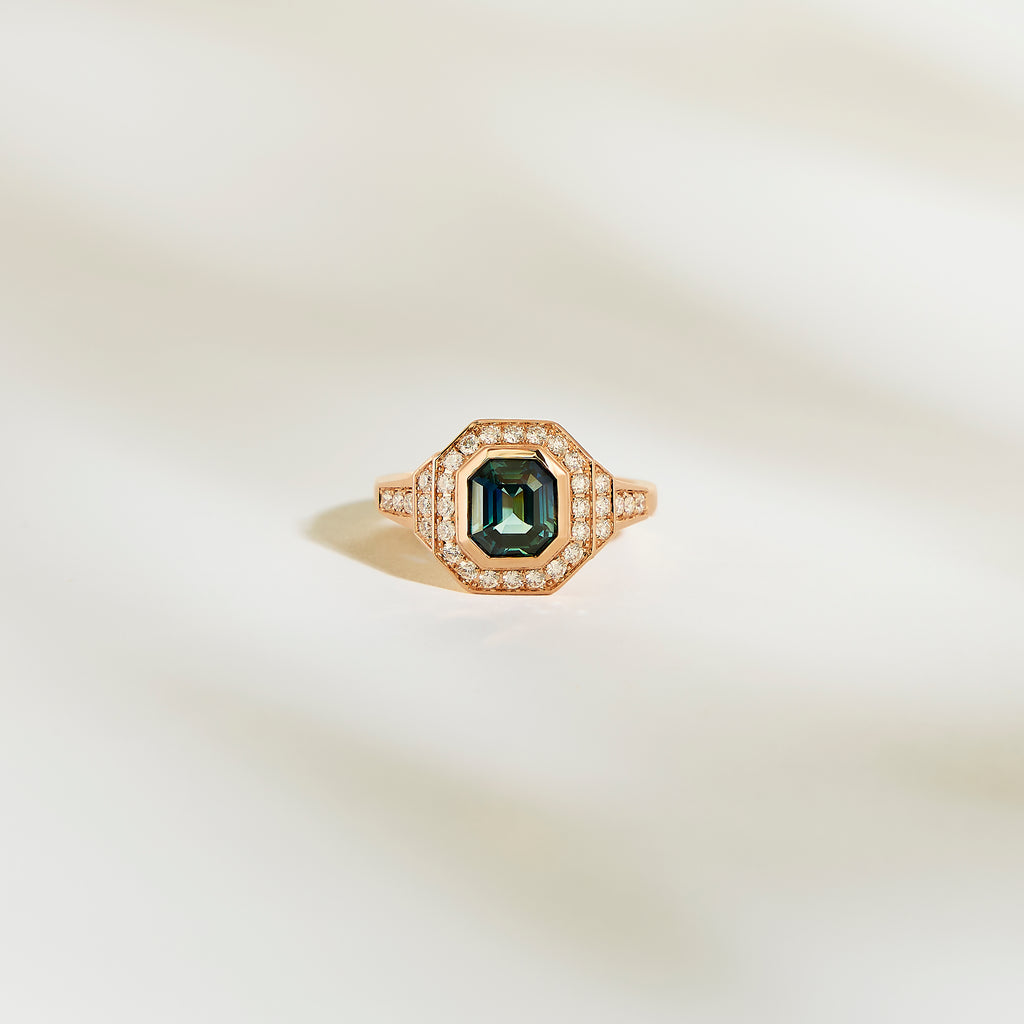 La Bague Saphir Teal de Bonnot Paris, une bague en or ornée d'une pierre précieuse vert foncé taille émeraude de 1,56 ct entourée d'un halo de petits diamants blancs, est élégamment mise en valeur sur un fond doux et clair.