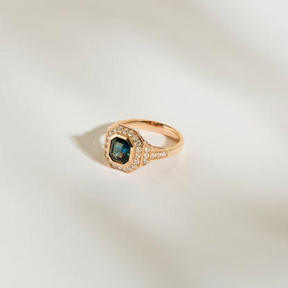 La Bague Saphir Teal de 1,56 carat signée Bonnot Paris arbore un saphir bleu-vert octogonal central entouré de petits diamants blancs. Elle repose élégamment sur une surface lisse et pâle, dont les ombres douces accentuent le design.