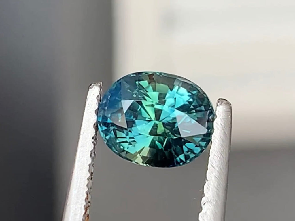 Une pierre précieuse ronde et facettée de la collection « Saphir Teal • 1,6 ct » de Bonnot Paris est maintenue par une pince métallique. Sa surface scintille de nuances de bleu et de touches de vert sur un fond neutre et flou.