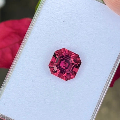 Une tourmaline rose hexagonale de 6,79 carats signée Bonnot Paris repose sur un fond blanc, ses facettes d'un rose éclatant étincelant. Une main la tient délicatement par le bord, avec un objet rouge flou en arrière-plan.