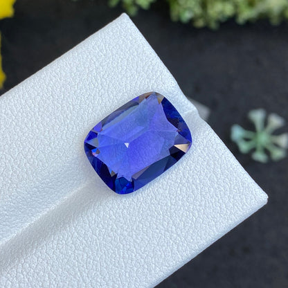 Un gros plan de la Tanzanite taille coussin de 6,66 ct de Bonnot Paris montre sa teinte bleue vibrante et ses facettes réfléchissantes, sur fond de surface texturée blanche avec des éléments verts flous en arrière-plan.