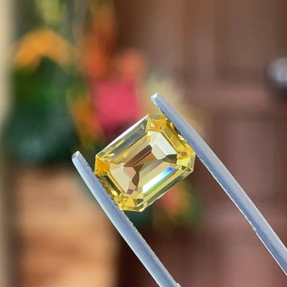 Une pierre précieuse jaune de 4,04 carats de la collection Saphir Jaune de Bonnot Paris est maintenue dans une pince métallique sur un fond flou de teintes vertes et brunes.