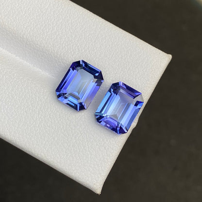 Deux tanzanites Bonnot Paris taille émeraude (5,82 ct chacune) sont placées côte à côte sur une surface blanche, présentant des couleurs vibrantes et des facettes précises sur un fond noir contrastant.