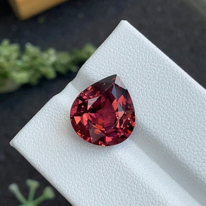 La Tourmaline Rose de 7,26 carats de Bonnot Paris est présentée sur une surface texturée gris clair, avec une plante verte floue en arrière-plan. Cette pierre précieuse rouge foncé en forme de poire reflète magnifiquement la lumière, mettant en valeur ses facettes et sa clarté.