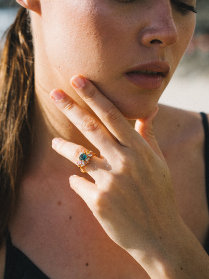 Une personne à la peau claire pose son menton sur sa main, mettant en valeur la bague Sea Bloom - Tourmaline de Bonnot Paris. Ses longs cheveux bruns sont attachés en arrière et elle porte un haut noir. L'image est doucement éclairée par la lumière naturelle, créant une atmosphère sereine.