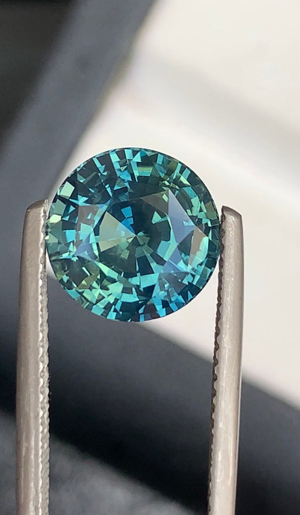 Une paire de pinces en métal maintient l'exquise pierre précieuse Bonnot Paris Saphir Teal • 5,52 ct sur un fond flou, où elle scintille dans des tons de turquoise et de sarcelle, soulignant sa taille complexe et sa clarté.