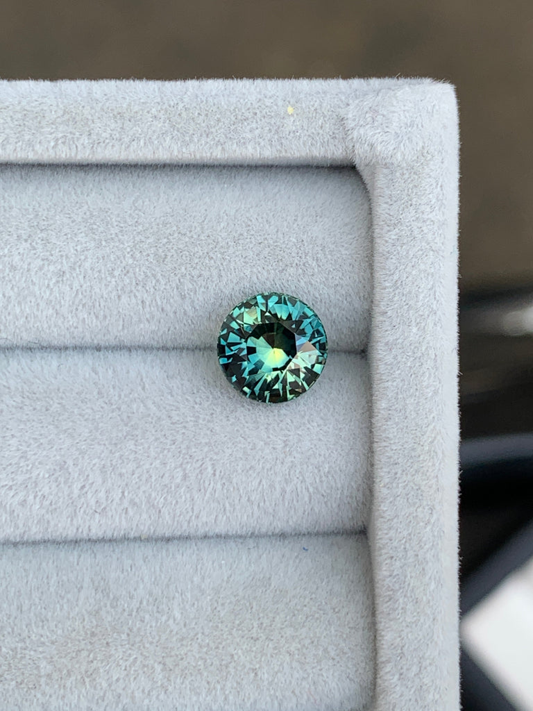 La pierre précieuse Bonnot Paris Saphir Teal • 2,55 carats de Madagascar scintille de reflets bleu sarcelle et aqua sur un fond gris doux, sa surface facettée évoquant l'élégance d'une exquise bague de fiançailles.