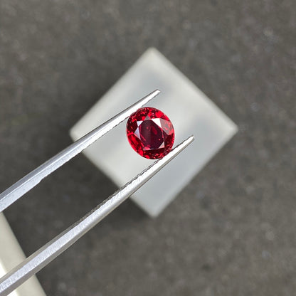 Une pince métallique maintient une pierre précieuse ronde facettée Spinelle Rouge · 1,25 carat de Bonnot Paris sur une surface texturée gris foncé. La pierre scintille de reflets lumineux, et un carré blanc flou apparaît en arrière-plan.