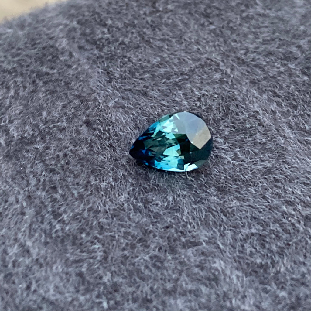 Un Saphir Teal en forme de larme de Bonnot Paris, pesant 2,06 carats, repose sur un tissu gris texturé, son allure non chauffée captivant tous ceux qui le contemplent.