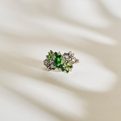 La bague Bonnot Paris Green Bloom - Tsavorite met en valeur une imposante pierre verte, entourée de saphirs bleus plus petits, de pierres incolores et de grenat tsavorite. Elle est élégamment présentée sur une surface crème doucement éclairée, projetant de subtiles ombres en arrière-plan.