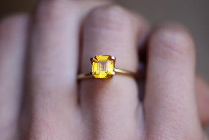 Une main met en valeur la bague Saphir Jaune · 2,04 ct de Bonnot Paris, comportant une pierre précieuse jaune taille carrée proéminente sertie d'or sur un fond flou.