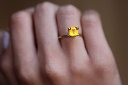 Une main portant la bague en or Saphir Jaune · 2,04 carats de Bonnot Paris avec une pierre précieuse jaune rectangulaire facettée sur un fond flou, soulignant la couleur vibrante de la bague et son sertissage élégant.