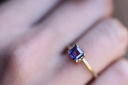Gros plan d'une main portant une bague en or Saphir Violet Bonnot Paris avec une pierre précieuse taille carrée de 1,65 carat, sertie sur un fond légèrement flou qui met en valeur les superbes détails de la bague.