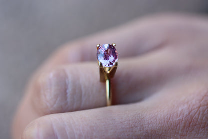 Une main ornée de la bague Saphir Violet · 1,61 carat en or de Bonnot Paris, mettant en valeur une pierre précieuse ronde et rose, est positionnée entre les doigts sur un fond légèrement flou.