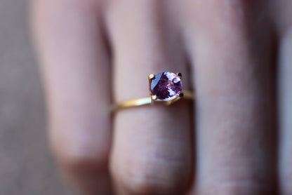 Un gros plan met en valeur une main portant la bague Saphir Violet en or 1,61 carat de Bonnot Paris, ornée d'une pierre violette à la taille scintillante, maintenue par quatre griffes. Elle se détache sur l'arrière-plan flou d'un doigt orné d'ongles neutres.