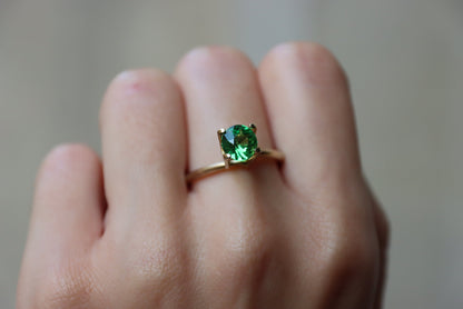 Une main présente une bague en or Bonnot Paris ornée d'une grosse pierre précieuse ronde Grenat Tsavorite de 1,49 ct sur l'annulaire, sur fond flou.
