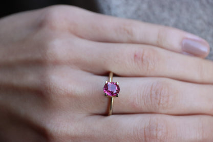 Une main ornée d'une bague en or 1,68 carat Rubis de Birmanie de Bonnot Paris, ornée d'une pierre rose ovale, repose délicatement sur un fond de tissu gris. Les ongles sont soigneusement manucurés avec un vernis naturel clair.