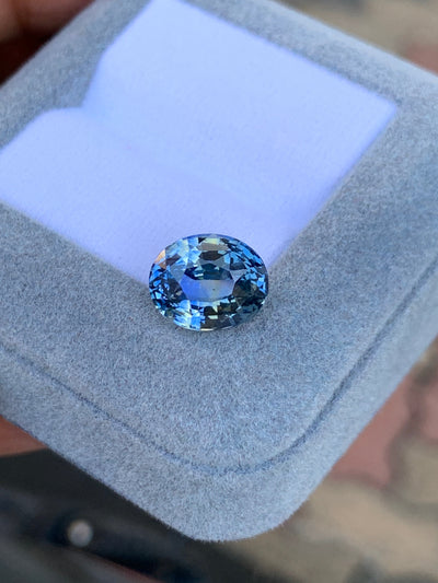 Le Saphir Antique • 6,2ct de Bonnot Paris, une pierre précieuse bleue taille ovale, repose sur une boîte à bijoux coussinée bleu clair avec un fond flou.