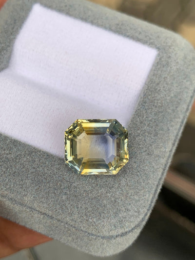 Le Saphir Antique taille émeraude (9,03 ct) de Bonnot Paris, aux superbes teintes jaunes et bleues, repose sur un couvercle de boîte à bijoux en velours gris clair, incarnant l'attrait des pierres précieuses uniques avec ses facettes magnifiquement inclinées et sa clarté.