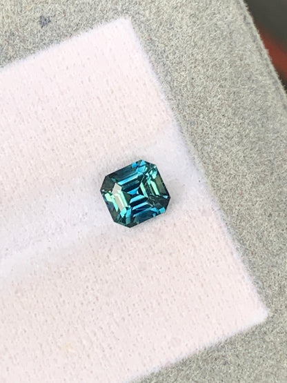 La [NOUVELLE] pierre précieuse Bonnot Paris Saphir Teal • 1,46 ct, de taille rectangulaire avec une teinte bleu-vert saisissante provenant des célèbres pierres précieuses de Madagascar, est présentée sur un fond texturé blanc et gris, mettant en valeur ses couleurs vibrantes et sa clarté à travers ses facettes réfléchissantes.