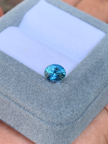 L'exquis Saphir Teal de 1,56 carat de Bonnot Paris, une pierre précieuse rare de Madagascar, est présenté dans un écrin gris ouvert doublé de tissu blanc. Cette pierre bleue ronde scintille de mille feux, mettant en valeur sa couleur vive et ses facettes saisissantes.