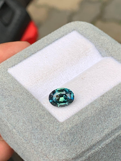 La pierre précieuse Saphir Teal 2,03 ct, de [NOUVEAU] Bonnot Paris, présente des teintes bleu et vert profonds, élégamment serties dans un coffret à bijoux gris avec une doublure blanche immaculée.