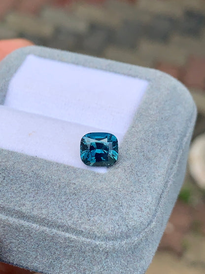 Le Saphir Teal • 2,50 carats de Bonnot Paris, un saphir taille coussin de 2,50 carats, est élégamment présenté dans un écrin de velours gris clair. Un fond légèrement flou met en valeur les détails exquis de ce trésor de Madagascar sur une surface aux motifs subtils.