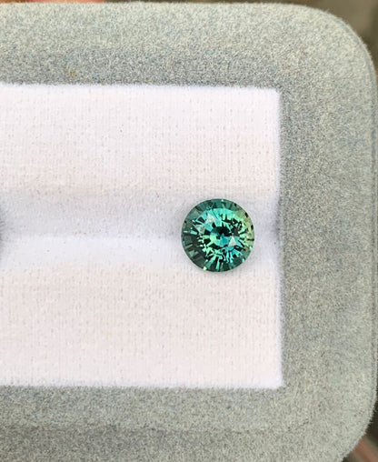 Une pierre précieuse Saphir Teal étincelante de 2,52 ct de [NOUVEAU] Bonnot Paris de Madagascar repose sur une plate-forme coussinée blanche et grise, affichant ses facettes bleu-vert vibrantes avec un fond doucement flou.