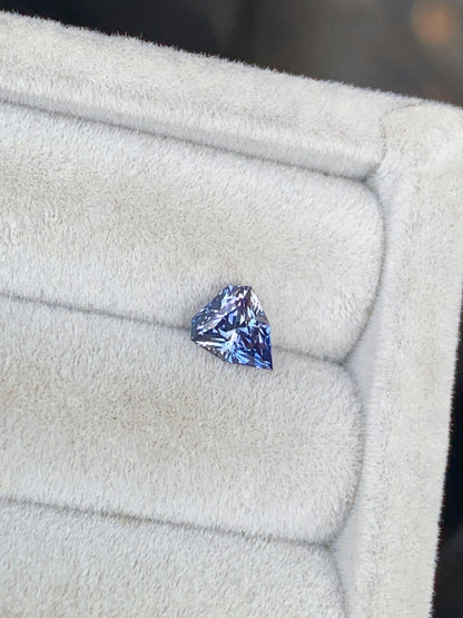 Un petit saphir bicolore triangulaire de 1,15 ct de Bonnot Paris, présentant des touches de violet, est élégamment positionné légèrement à gauche sur un plateau à bijoux texturé gris clair, illustrant la joaillerie d'exception.
