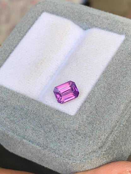 Le Saphir bicolore • 1,51 ct de [NOUVEAU] Bonnot Paris est une pierre précieuse violette taille émeraude éclatante, posée sur un coussin blanc dans un écrin gris clair. Ses facettes éclatantes et polies sont magnifiquement mises en valeur par des reflets.