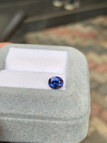 Un saphir bleu de 1,25 carat, taille ovale et éclatant, repose sur un tissu blanc dans un écrin gris ouvert signé Bonnot Paris. Les carreaux texturés et flous en arrière-plan rehaussent l'attrait de ce joyau exquis de Madagascar.