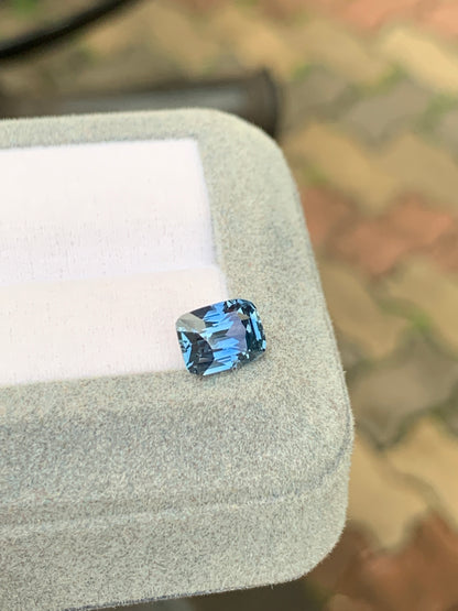 Un Saphir bleu de 2,75 carats de Bonnot Paris, taille coussin, est présenté dans un écrin gris clair. Le fond brun et vert délicatement flouté met en valeur cette pierre précieuse rare.