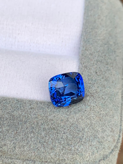 Un saphir bleu éclatant de 3,02 carats de Bonnot Paris, taille coussin, repose sur une surface grise douce sur fond blanc. Cette pierre précieuse exquise reflète magnifiquement la lumière et crée de superbes motifs.