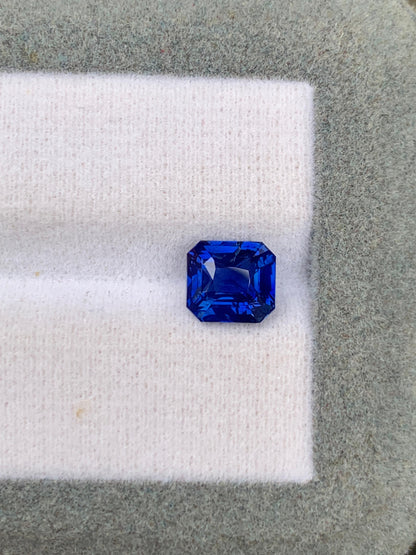 Un saphir bleu taille émeraude d'un bleu profond de 2,53 carats, provenant du Sri Lanka, signé [NOUVEAU] Bonnot Paris, repose sur un fond de tissu blanc et vert, mettant en valeur ses facettes brillantes et sa couleur intense. Cette pierre précieuse non chauffée séduit par son charme naturel.