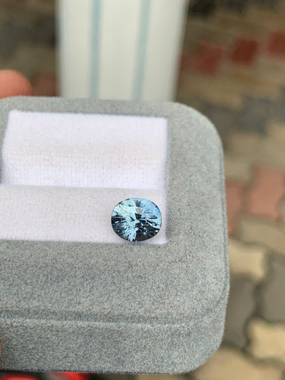 Un saphir gris bleu rond de 3,10 carats de Madagascar de Bonnot Paris scintille sur une base coussinée blanche à l'intérieur d'un coffret en velours gris, avec des pavés terreux flous en arrière-plan pour souligner son élégance.