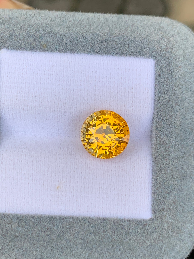 Un saphir jaune taille brillant rond de 5,75 ct de Bonnot Paris scintille de couleurs vibrantes et de facettes complexes sur un fond coussiné blanc et gris, incarnant l'essence d'un bijou unique.