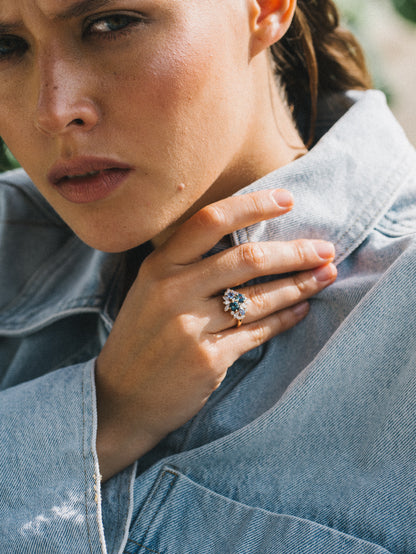 Vêtue d'une veste en jean légère, quelqu'un touche sa poitrine pour révéler la bague Bonnot Paris "Teal Bloom - Saphir", comportant une pierre précieuse verte frappante entourée de diamants scintillants sur un fond légèrement flou.