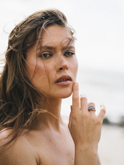 Une personne aux longs cheveux bruns se tient dehors, regardant droit vers l'objectif. Elle se touche le menton, mettant en valeur une bague colorée Indigo Bloom - Saphir de Bonnot Paris. L'arrière-plan présente une scène naturelle floue.