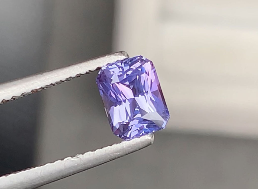 Une pince à épiler tient un saphir violet étincelant de 1,06 carat, taille rectangulaire, de la marque Bonnot Paris. Ses teintes violettes vibrantes, proches de celles des pierres précieuses du Sri Lanka, reflètent la lumière à travers ses multiples facettes pour un effet éblouissant sur un fond flou.