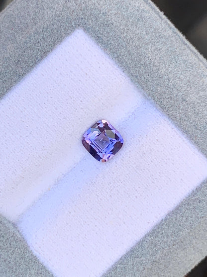 Un saphir violet taille coussin de 1,08 ct de chez Bonnot Paris est posé au centre d'un écrin gris clair doublé de tissu blanc. La pierre scintille sous la lumière, accentuant sa couleur vive et ses facettes.