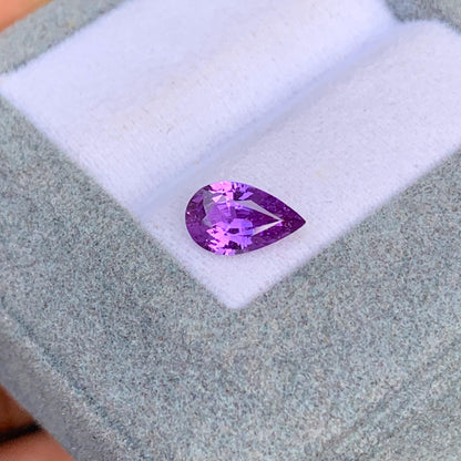 Une pierre précieuse violette Saphir (1,18 ct) de [NOUVEAU] Bonnot Paris repose sur un tissu gris clair dans une boîte à bijoux ouverte, sa couleur vibrante et ses facettes brillant de clarté et de qualité rappelant les célèbres pierres précieuses du Sri Lanka.