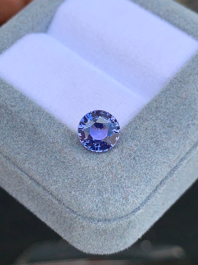 Le Saphir violet rond facetté • 2,45ct de [NOUVEAU] Bonnot Paris scintille sur un coussin blanc à l'intérieur d'une boîte gris clair, mettant en valeur sa clarté et sa taille complexe parmi d'autres pierres précieuses.