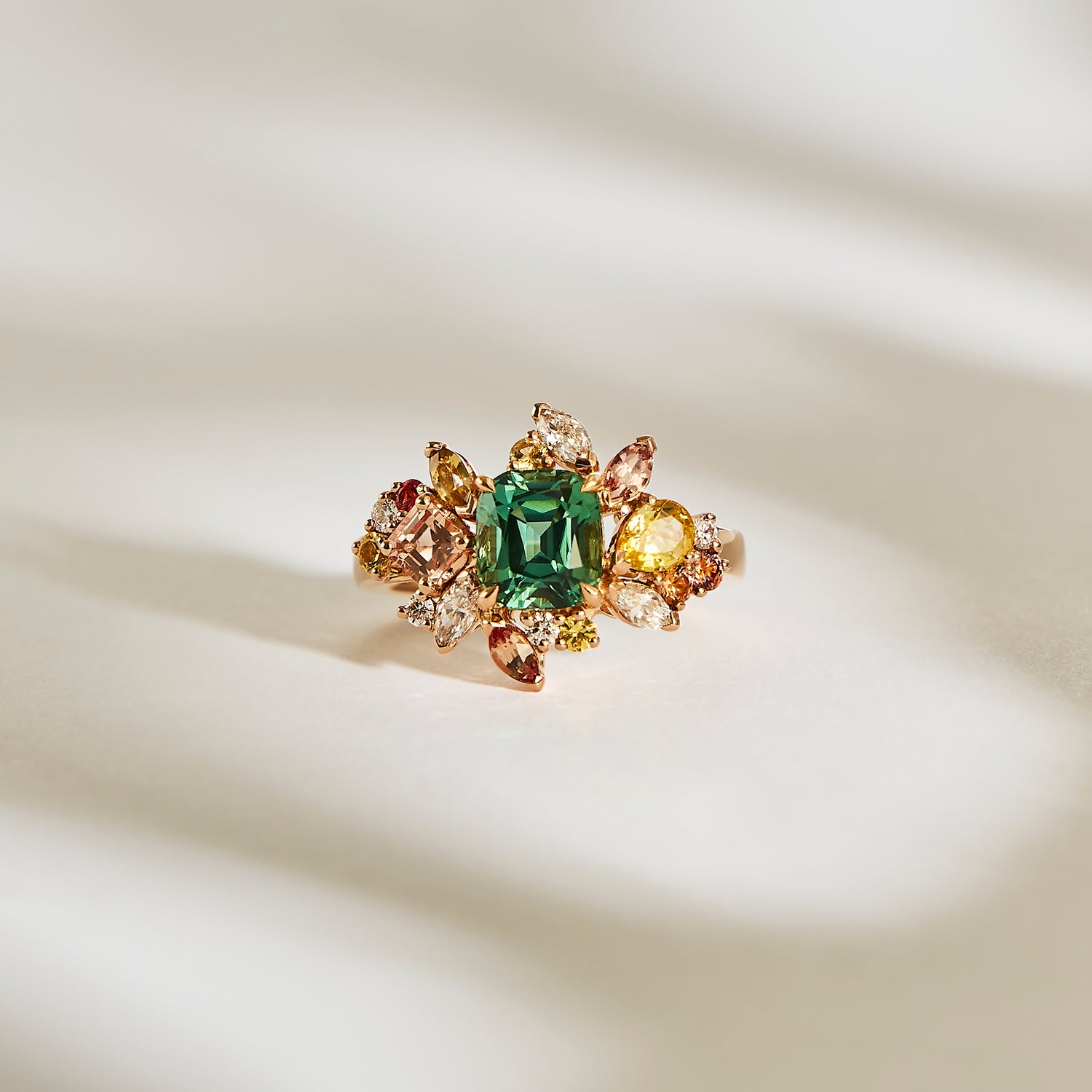 La Sea Bloom - Tourmaline de Bonnot Paris est une bague en or ornée d'une grosse pierre précieuse verte au centre, entourée de saphirs champagne et de diamants dans un motif floral, magnifiquement présentée sur une surface blanche texturée et doucement éclairée.