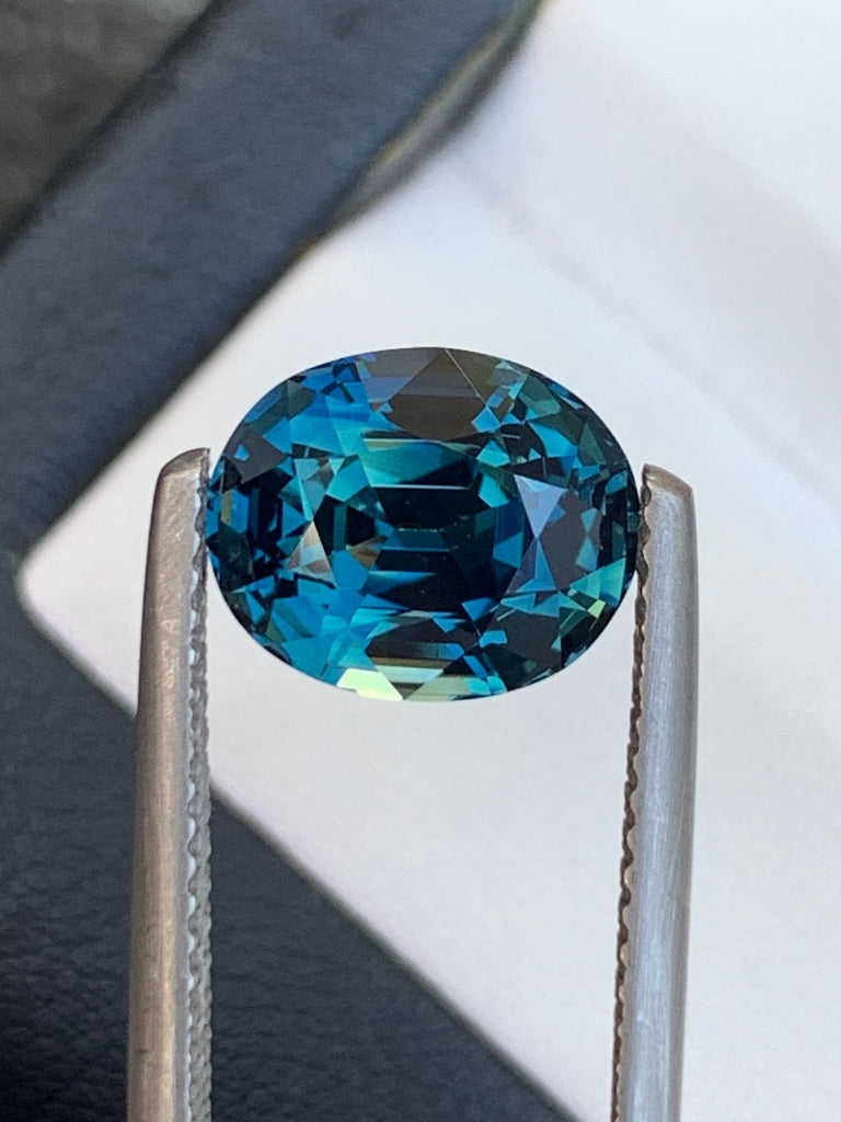 Gros plan de la pierre précieuse Saphir Teal de Bonnot Paris de 4,1 carats, photographiée à la pince à épiler sur un fond flou. Cette pierre ovale présente des facettes complexes qui reflètent des nuances fascinantes de bleu et de vert, idéales pour une bague de fiançailles saisissante.