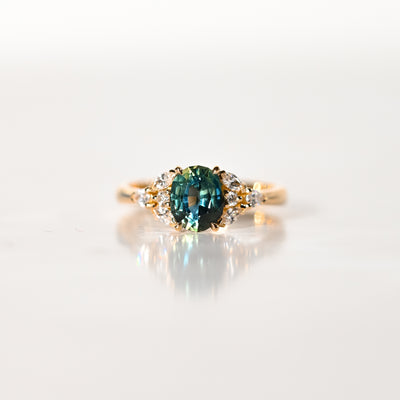 La Bague Saphir Teal · 1,5 carat de Bonnot Paris [NOUVEAU] est ornée d'un saphir ovale turquoise saisissant, encadré de diamants blancs ronds. Elle repose élégamment sur une surface blanche lisse et réfléchissante, telle une bague de fiançailles raffinée.