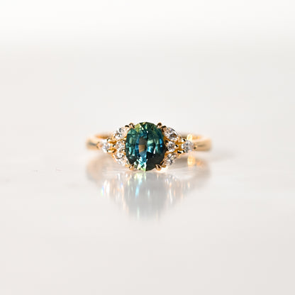 La Bague Saphir Teal · 1,5 carat de Bonnot Paris [NOUVEAU] est ornée d'un saphir ovale turquoise saisissant, encadré de diamants blancs ronds. Elle repose élégamment sur une surface blanche lisse et réfléchissante, telle une bague de fiançailles raffinée.